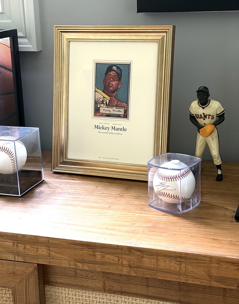A framed Collector&rsquo;s Story print of the 1952 Topps Mickey Mantle in a gold frame on a wooden sideboard, beside two signed baseballs